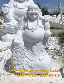 Tượng Phật Di Lặc đứng cầm súng như ý màu trắng ngồi trên một đống đá.