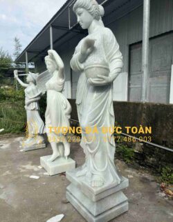 A TƯỢNG CÔ GÁI ĐÁ CẨM THẠCH TRẮNG NGUYÊN KHỐI ĐẸP TỰ NHIÊN cầm giỏ trước tòa nhà.