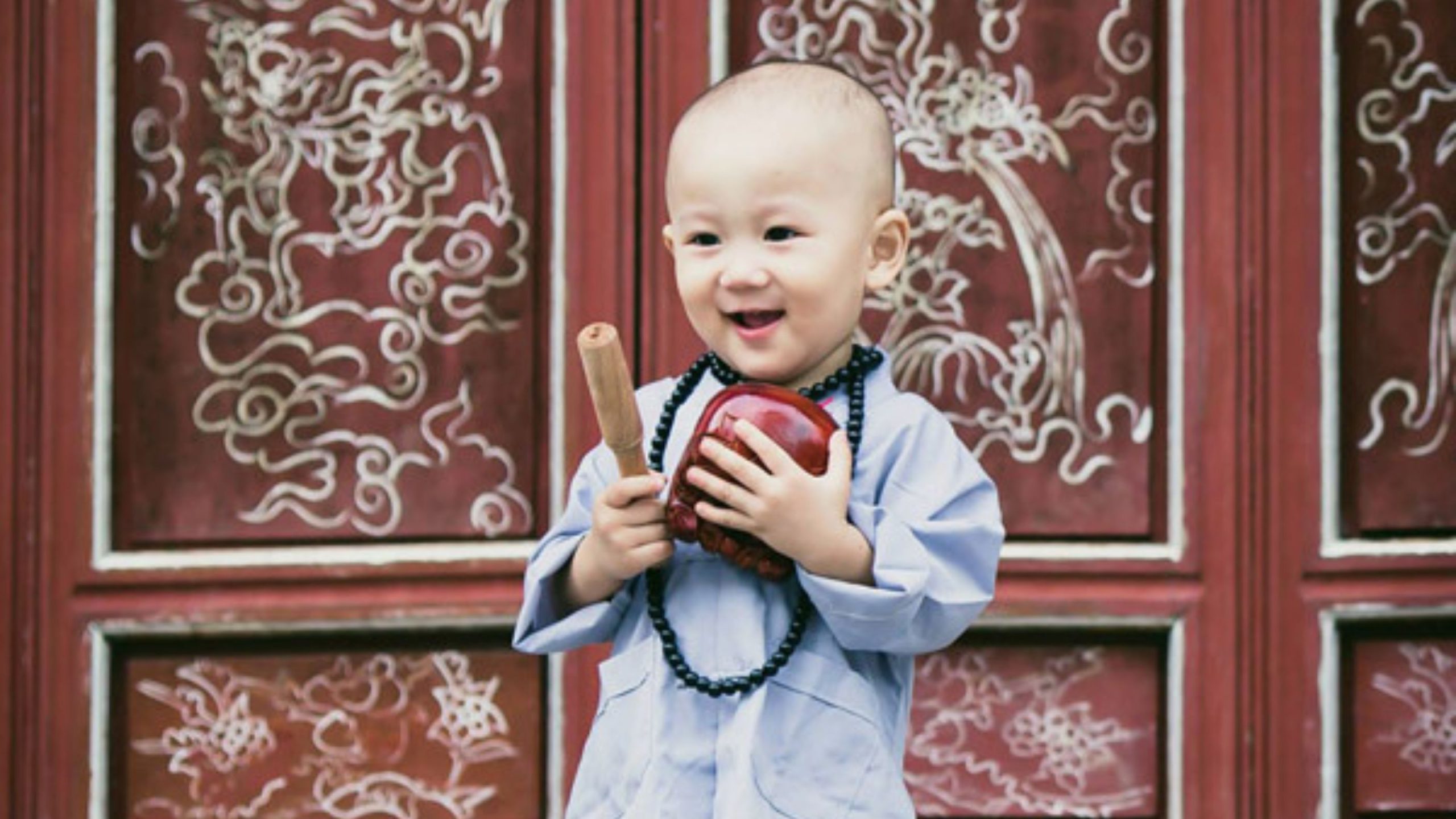 Chú tiểu cầm nhạc cụ trước cánh cửa màu đỏ.